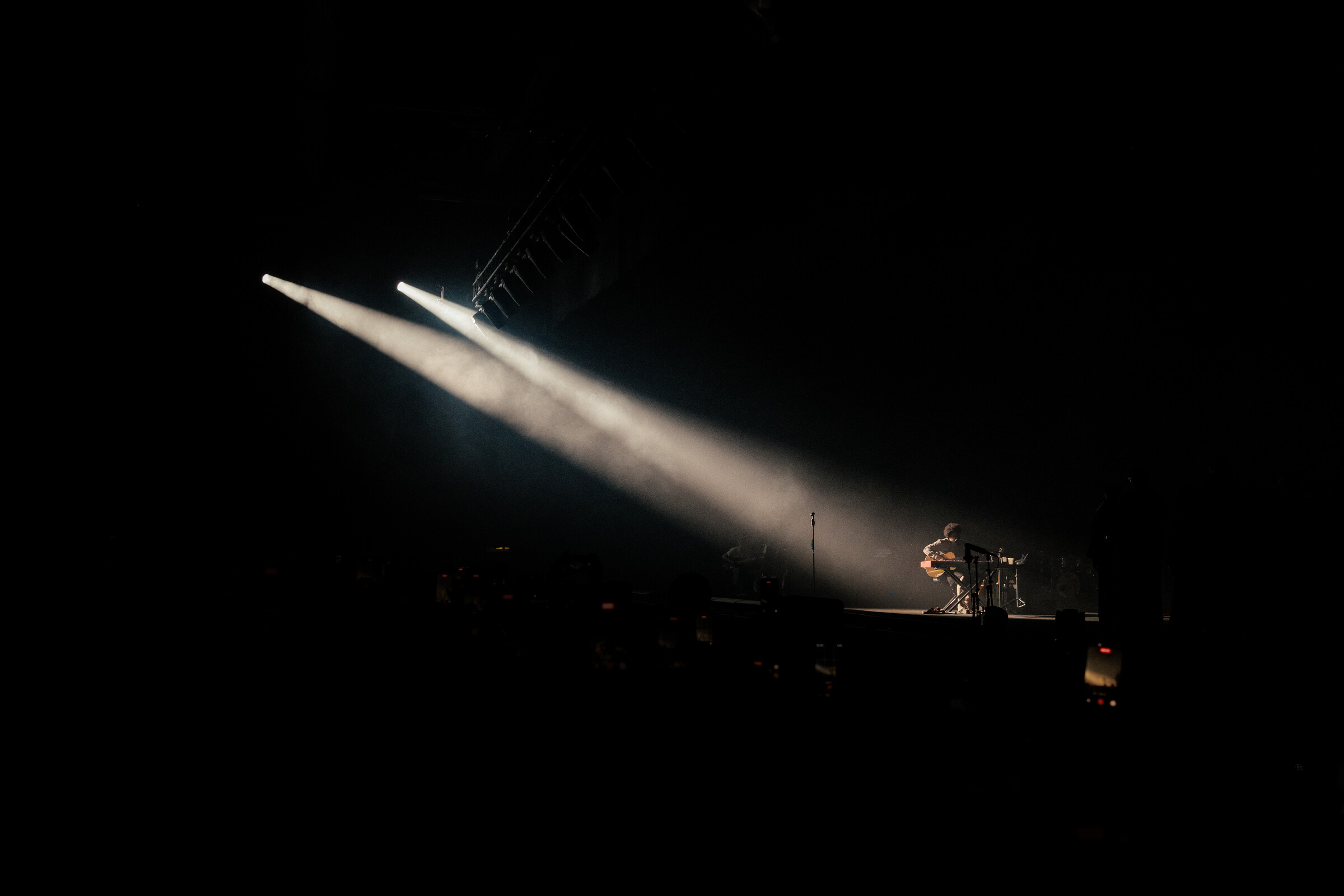 A single small figure with a guitar under two crossed spotlight beams, dwarfed by dark stage space